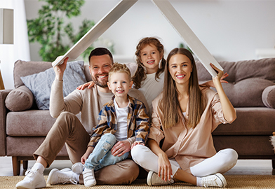 Family of four sitting in front of couch holding cardboard home roof