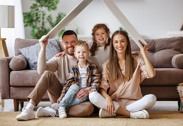 Family of four sitting in front of couch holding cardboard home roof